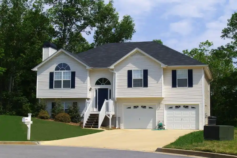 A wide shot of a classic two-story suburban house with white siding, navy blue shutters, a two-car garage, and a well-manicured green lawn under a blue sky.