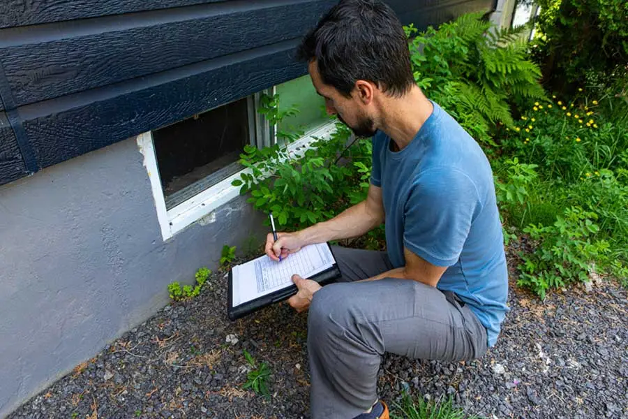 A pest control professional in a blue shirt kneels outside a house, filling out a Wood Destroying Insect (WDI) inspection report on a clipboard near the home's foundation.