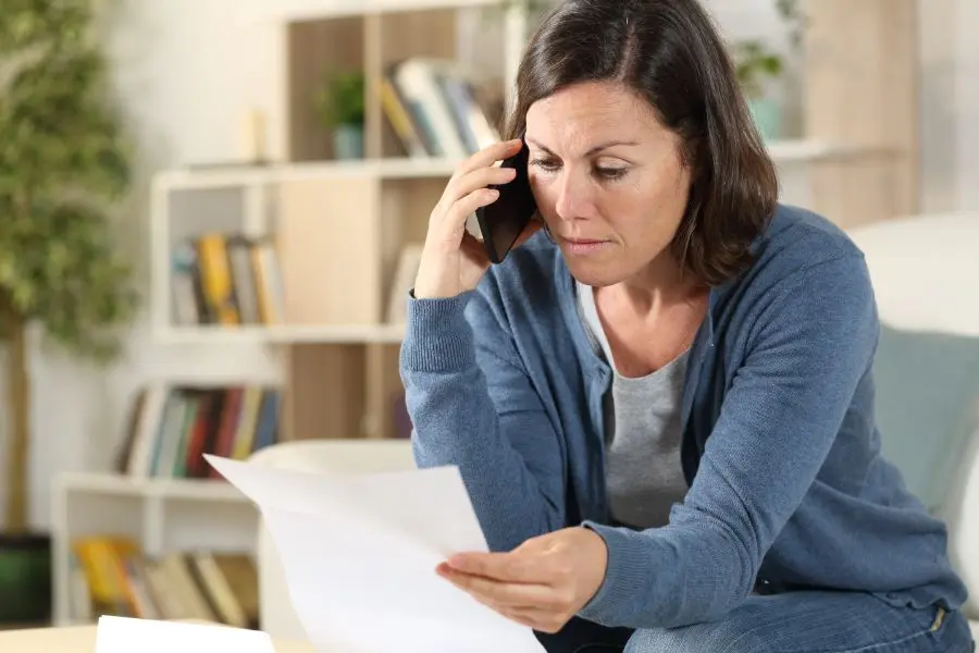 A woman looking concerned and stressed while sitting in her living room, holding a white document in one hand and talking on a smartphone with the other.
